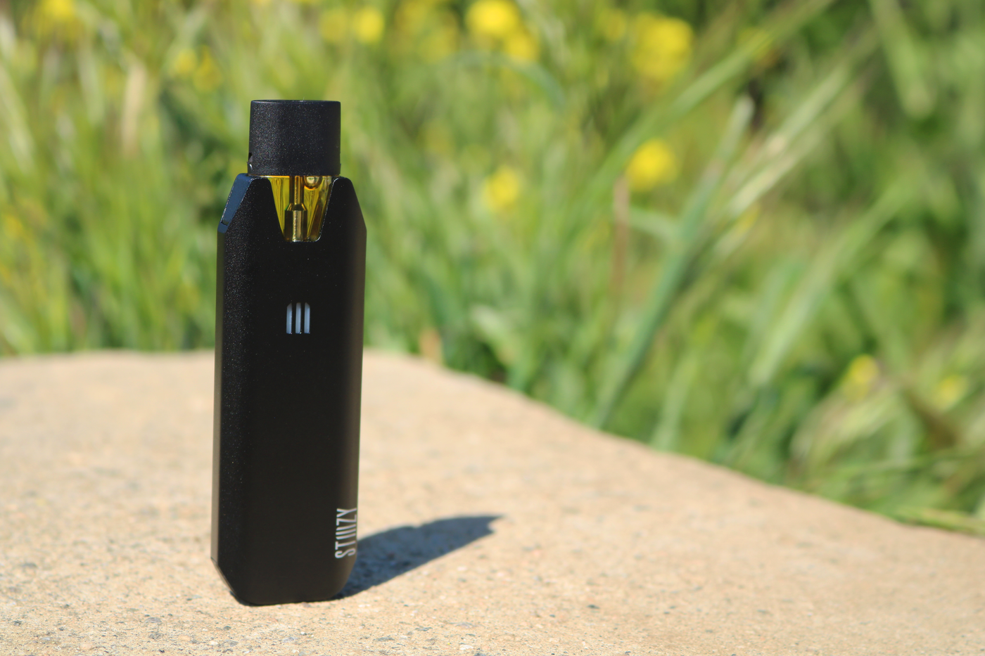 A black vape pen for cannabis from STIIIZY with a big battery overlooks a lawn.